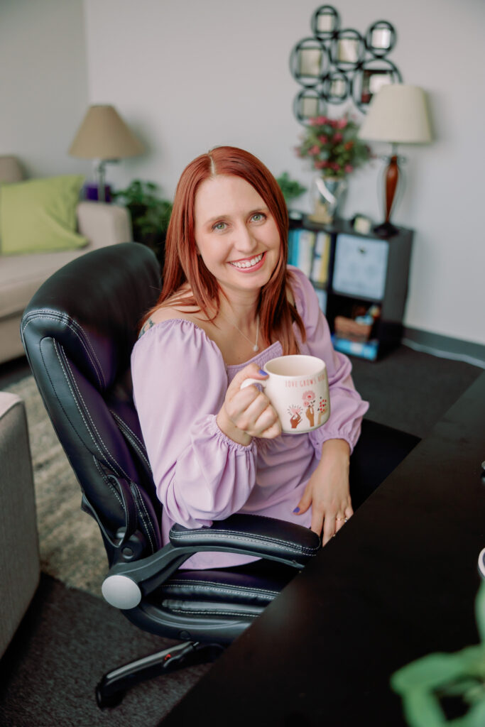 The therapist is smiling and sitting in a chair in her office holding a cup of coffee.